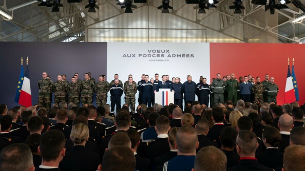 Le président de la République derriè-re un embon, de part et d'autres de nombreux militaires de toutes les armées. A arrière-plan, un fond bleu-blanc-rouge et à gauche et à droite les drapeaux français et européen