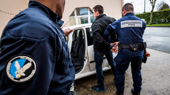 Deux gendarmes de PSIG en combinaison bleue marine en train d'arrêter un individu vêtu d'un jean et blouson noir devant un véhicule blanc