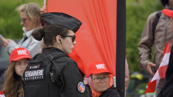 une gendarme mobile de trois quart arrière droite devant des presonnes coiffées d'une casquette rouge de la Haute-Savoie et devant un drapeau aux mêmes couleurs