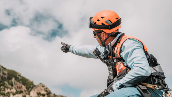 Un gendarme sur un rocher, portant une chasuble "secours en montagne", regardant vers les hauteurs et les désignant de la main droite
