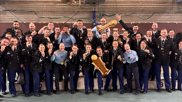 Une trentaine de gendarmes, hommes et femmes, dont la majorité est en vareuse et les autres en tenue d'intervention, posent en groupe dans un gymnase, sur et devant un podium. Au milieu, deux gendarmes brandissent chacun un trophée. Tous ont une médaille autour du cou.