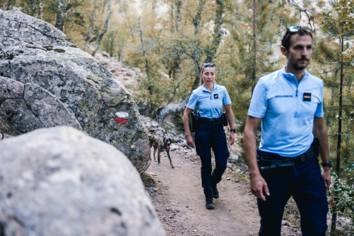 Deux gendarmes (un homme et une femme) évoluent sur un sentier de randonnée, l'un derrière l'autre. Derrière eux se tient un chien marron de taille moyenne.