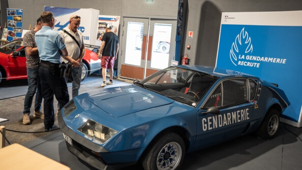 Une Alpine bleue de gendarmerie ancien modèle exposée, devant un panneau bleu "la gendarmerie recrute". A gauche, un gendarme échange avec deux civils