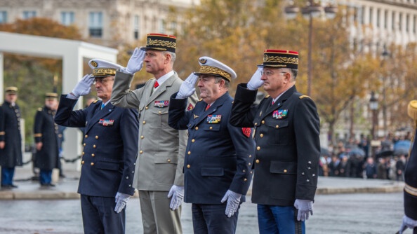 Quatre militaires, trois en tenue sombre, le quatrième vêtu d'une tenue beige, se tiennent deboit côte à côte. Leur bras gauche est positionné le long de leur cuisse gauche, tandis qu'ils effectuent un salut de la main droite. Derrière eux, on apercçoit des arbres dont les feuilles sont aux couleurs de l'automne, et la façade de bâtiments en pierre.