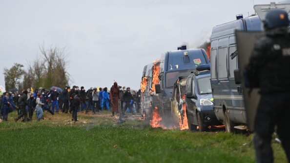 Une groupe d'opposants aux bassines encagoulés et armés au second plan. Au premier plan, des véhicules de gendarmerie en feu.