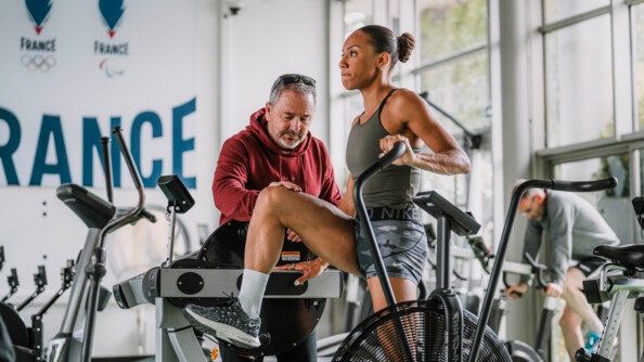 Une sportive vêtue d'une tenue grise, effectue un exercice sur un appareil dans une salle d'entraînement physique. Son entraîneur, qui se tient debout à ses côtés, accompagne le geste en posant sa main sur son genou. La photo est prise de biais. Derrière, en gros sur le mur, est écrit le mot FRANCE