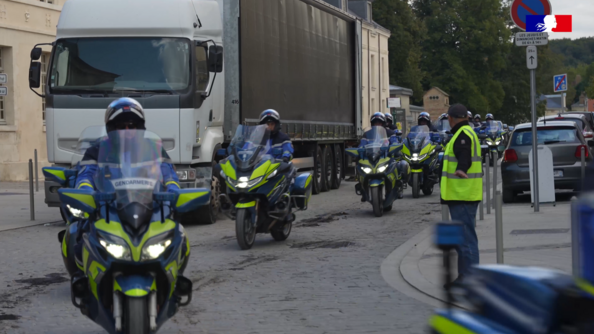 Des gendarmes motocyclistes circulent en file, dans une rue pavée. Les militaires longent un gros camion stationné le long du trottoir. Un homme, vêtu d'un gilet de sécurité jaune, assiste au passage du convoi.