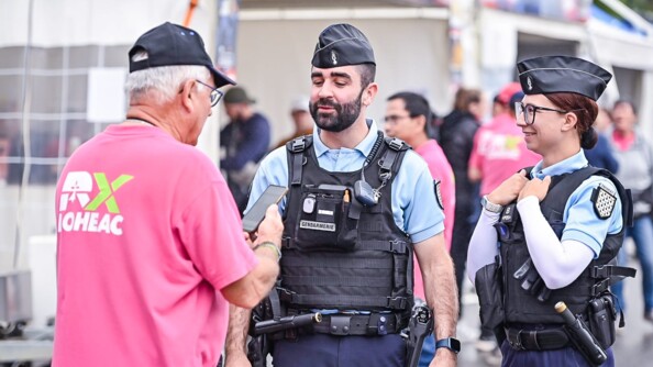 Deux gendarmes, une femme à droite et un homme à gauche, discutent avec un homme âgé de dos, porteur d'une casquette noire et d'un polo rose floqué "RX LOHEAC", avec un téléphone dans la main droite