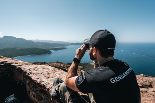 Un gendarme, assis sur un muret en pierres surplombant la mer, regarde l'horizon avec des jumelles. Le militaire se tient de dos et l'on peut lire dan sle dos de son polo noir l'inscription " Gendarmerie". il porte aussi une casquette noire et unpantalon treillis kaki. On aperçoit les montagnes bordant la mer.