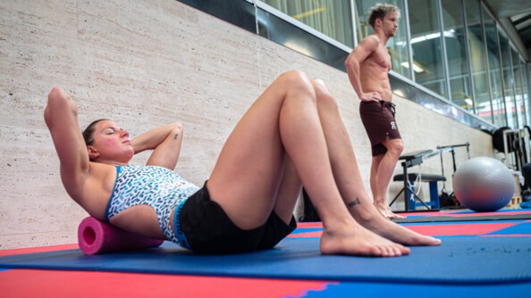 Une sportive vêtue d'un maillot de bain blanc et bleu, ainsi que d'un short noir court, réalise des exercices d'étirement, allongée sur le dos, les yeux fermés, sur un tapis de sol de couleur bleue. Sa nuque repose sur un appui tête rose.