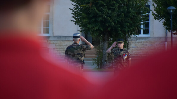 Au premier plan, vue floue sur les épaules de deux sportifs vêtus de rouge. Au centre de la photo, la netteté est faite sur deux officiers de gendarmerie en treillis qui se saluent.