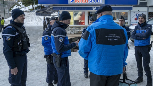 Devant un intersport et sur la terre enneigée, de part et d'autre du'n homme à la doudoune bleue, de dos, siglée "Gourette La Pierre Saint Martin", six gendarmes