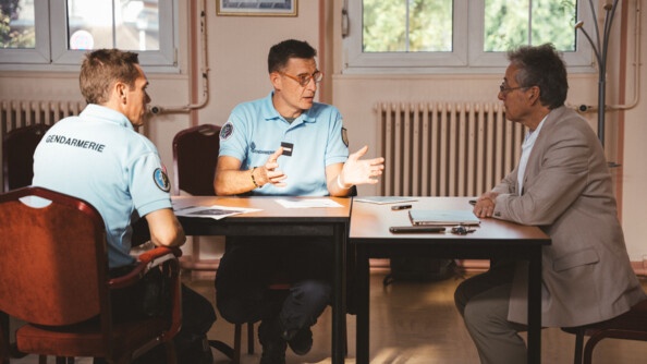 Deux gendarmes et un hommes en costard beige autour d'une table recouverte de documents et d'un ordinateur portable