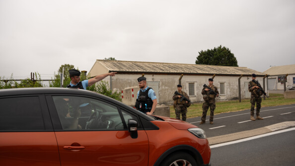 Au premier plan, un véhicule couleur rouille, et au second plan, deux gendarmes, dont l'un montre une direction au conducteur. En arrière plan, sur le bord de la route, trois militaires de Sentinelle armés de fusils. Derrière eux, une clôture haute avec des barbelés devant des baraquements.