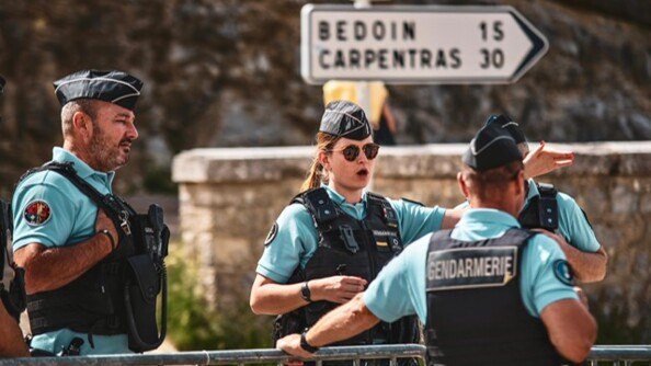 Quatre gendarmes, un de profil, une femme de face, en train de parler et d'indiquer une direction, un homme de dos, la main contre une barrière, et un quatrière que l'on devine. Les quatre militaires sont sur le bord d'une route dans le Vaucluse.