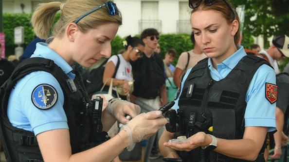Deux jeunes femmes gendarmes se tiennent debout, l'une en face de l'autre. Les deux militaires tiennent en main leur téléphone portable professionnel. Et l'une tient une petite coupelle ronde transparente posée à plat dans sa main gauche. La scène se déroule à l'occasion du festival Garorock