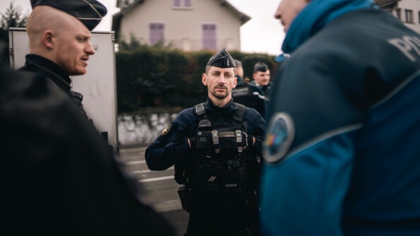 Deux gendarmes discutent avec un policier suisse. La scène se déroule en éctérieur, le long d'un avenue, par temps gris. On aperçoit la façcade d'une maison beige aux volets mauves, en second plan, de l'autre côté de la rue.