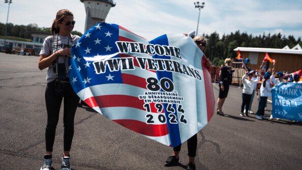 Deux femmes sur un tarmac tiennet un grand drapeau américain en forme de coeur surmonté des inscriptions en noir "WELCOME WWII VETERANS 80e anniversaire du débarquement en Normandie 1944 2024"