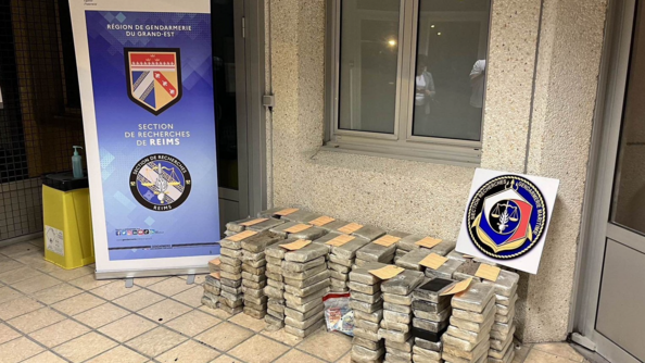 Des dizaines de paquets rectangulaires contenant de la cocaïne sont posés sur le sol, avec un sac de scellés contenant du numéraire. Un panneau portant l'écusson de la S.R. apparaît à droite, tandis qu'un kakémono de la région de gendarmerie Grand-Est et de la S.R. de Reims est posé à gauche.