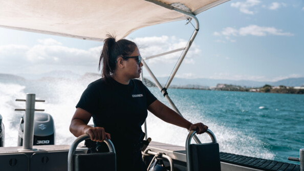 Une femme gendarme, vêtue d'une tenue noire, et de lunettes de soleil noires, se tient debout sur une embarcation, évoluant sur la mer. On aperçoit de nombreuses projections à l'arrière du bateau, liées à la vitesse, ainsi que les reliefs de la côte. La femme se tient des deux mains aux deux sièges situés devant elle.
