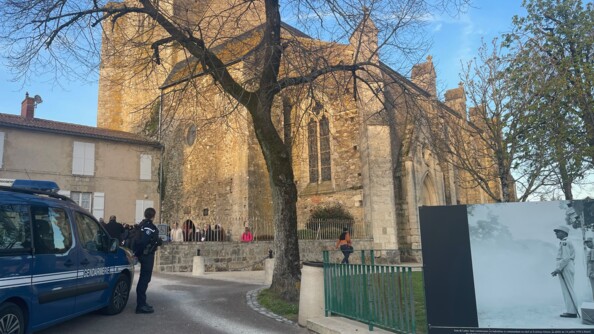 à gauche un véhicule gendarmerie, devant lui une gendarme de profil. au premier plan à droite une photo de Jean de Lattre devant un parc d'herbe. Au second lan au centre un arbre sans feuille. En arrière plan une église où se reflète le soleil