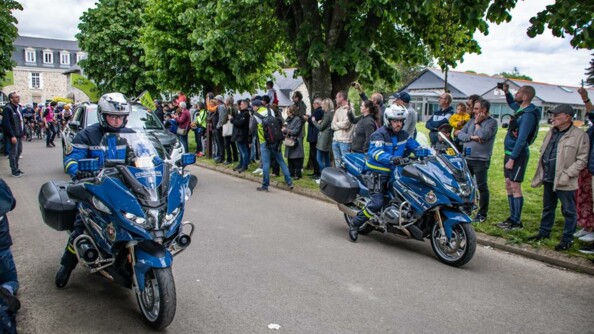 Deux motocyclistes de la garde républicaine de part et d'autre de la route, au bord de laquelle sont amassés des spectateurs.