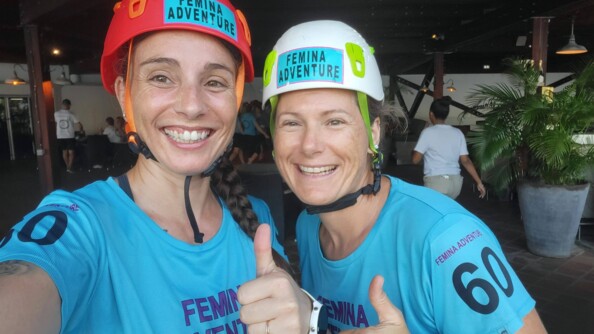 Deux femmes posent côte à côte, souriantes, le pouce levé. Elles sportent le moême t-shirt bleu turquoise avec l'inscription FEMINA ADVENTURE,  aini qu'un casque (rouge pour l'une, et blanc pour l'autre) portant aussi l'inscription FEMINA ADVENTURE.