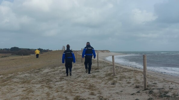 Deux gendarmes de dos, un homme et une femme, avancent en bordure de plage, sur un terrain sablonneux. A leur droite, se trouvent la plage et la mer à marée haute. Au loin devant eux, on aperçoit une personne vêtue d'un ciret jaune, qui avance, le corps courbé en avant. Le ciel est très gris.
