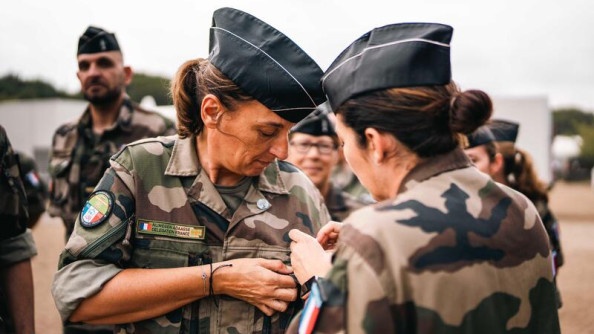 Deux femmes militaires se tiennent debout, l'une face à l'autre, en tenue kakie, avec un calot noir sur la tête. L'une accroche quelque chose, que l'on ne distingue pas, à la poitrine de la seconde militaire. Toutes deux portent au bras un écusson représentant le drapeau français.