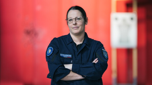 Une femme gendarme, brune, avec des lunettes, vêtue d'une tenue noire, se tient debout, face à l'objectif, bras croisés contre la poitrine, affichant un léger sourire. La scène se déroule dans une pièce aux murs rouges. On devine derrière elle une cible de tirs, dont l'image a été floutée.