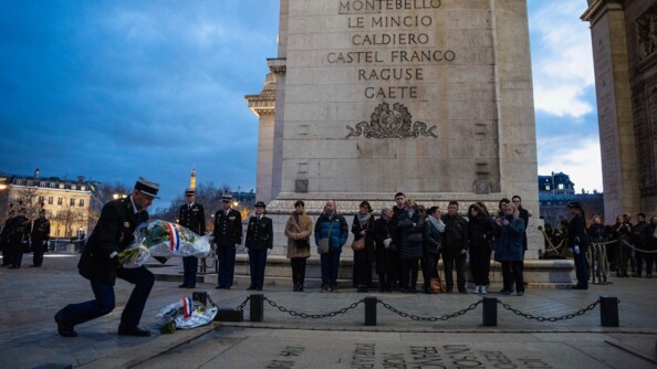 Sous l'Arc de Triomphe, un gendarme s'agenouille pour d&eacute;poser une gerbe de fleurs sur la tombe du soldat inconnu.  En arri&egrave;re plan, des personnes en tenue militaire et des civils.
