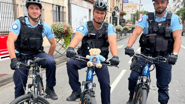 Trois élèves-gendarmes posent sur leur VTT, un pied à terre, face caméra, dans une rue de Paris. Un petit nounours revêtu d'un polo gendarmerie avec une rondache en son centre, est posé sur le cadre du vélo du milieu.