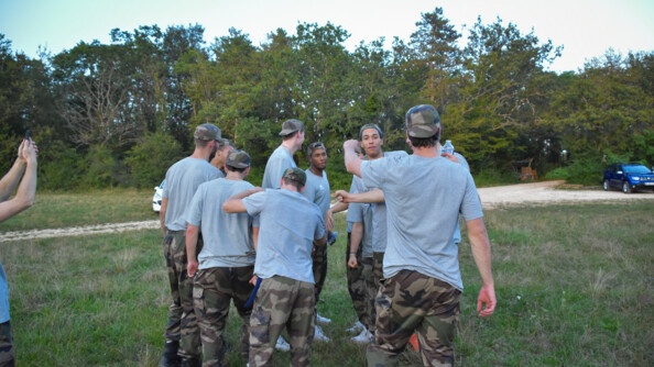 Au milieu d'un champ, en lisière de bois, un groupe de neuf jeunes hommes en bas de treillis kaki et tee-shirts gris, regroupés en train de faire un check collectif.