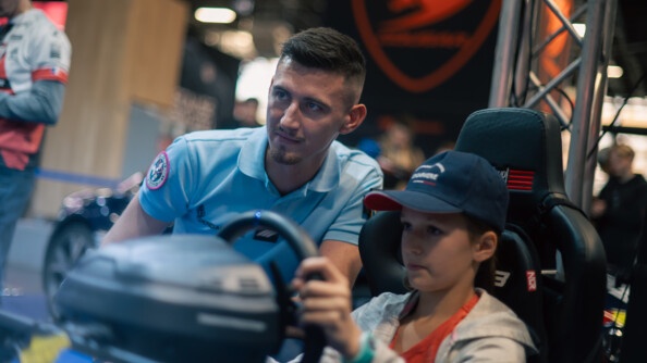 Un gendarme en tenue est accroupi aux côtés d'un jeune garçon au volant d'un simulateur de conduite. Le garçonnet porte une casquette bleue marine sur laquelle on devine l'inscription Gendarmerie.