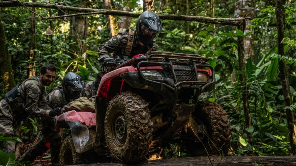 Militaire faisant passer son quad au dessus d'un tronc avec l'aide de deux camarades.