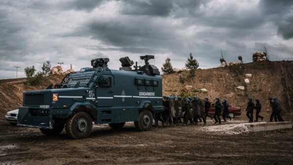Un peloton de gendarmerie mobile en colonne d'assaut derrière un Centaure sur un terrain boueux. Derrière eux, un talus