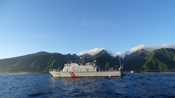 Photo d'un patrouilleur gris, avec sur la coque des bandes bleues, blanches et rouges, naviguant en bordure de côte, où se dressent des reliefs verdoyants, auxquels s'accrochent quelques nuages