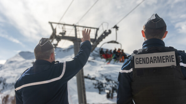Deux gendarmes se tiennent debout, dos à l'objectif, regardant en direction d'un télésiège sur lequel se trouvent des touristes. Le gendarme situé sur la gauche de l'image agit la main en direction des skieurs. Dans le ciel, le soleil perce au travers des nuages.