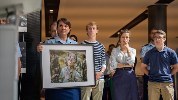 Pr&eacute;sentation de la photo de Christophe Fernandez qui a &eacute;t&eacute; choisi par le g&eacute;n&eacute;ral Christian Rodriguez. Derri&egrave;re elle, se tient des invit&eacute;s concentr&eacute;s sur la pr&eacute;sentation. La photo est un portrait d'une femme qui se fait maquiller le visage par un camarade pour &ecirc;tre camoufl&eacute;. Elle a le sourire et fait un clin d'&oelig;il.