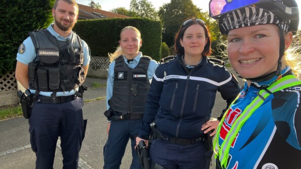 une jeune femme cycliste avec à sa gauche trois gendarmes (un homme et une femme). Tous les quatre sourient. Ils se trouvent dans la rue