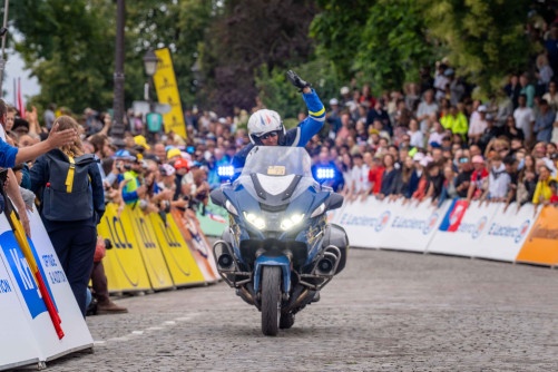 Photo d'un motocycliste de la Garde Républicaine, dans les rues de Paris, levant le bras gauche en direction de la foule massée sur le bord de la chaussée