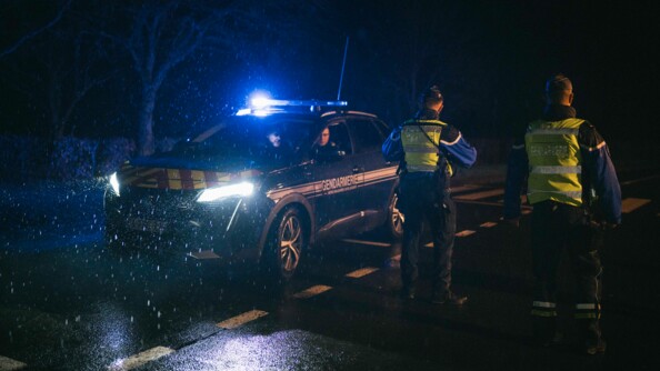 4 gendarmes de nuit sont sur la route pour un contrôle routier. Sur la droite, 2 d'entre eux sont debout de dos. Et sur la gauche, 2 autres sont dans un véhicule, phares allumés, sous la pluie.