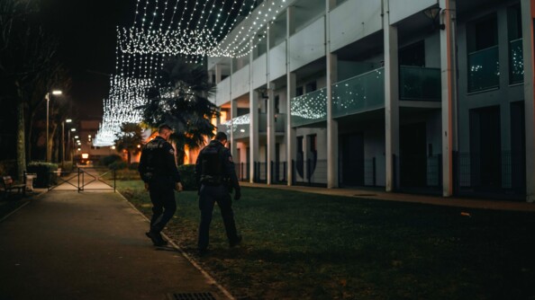 Deux gendarmes qui marchent au pied d'un immeuble. Derrière se trouve des guirlandes de Noel.