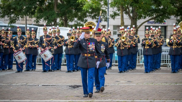 Des gardes de la garde républicaine donnent un concert debout sur un parvis, tandis que deux s'avancent à l'avat de l'image.