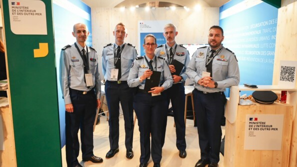 Au stand MIOM de l'ANCTour, cinq gendarmes en chemise bleue, cravate et pantalon droit, posent. A gauche, deux colonels. Au centre une adjudant-chef (ADC) référente sûreté, et son camarade ADC référent sûreté à droite, tenant tous les deux une tablette neogend. A droite, un autre adjudant-chef.