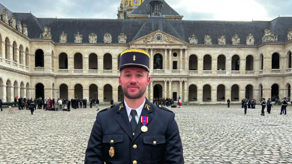 Un jeune homme brun, portant la barbe, en uniforme bleu marine et un képi bleu marine et doré, pose debout, les deux mains jointes devant lui, dans une cour pavée (il s'agit de la cour d'honneur des invalides). A l'arrière, on aperçoit le dôme des invalides, ainsi que le ciel gris.