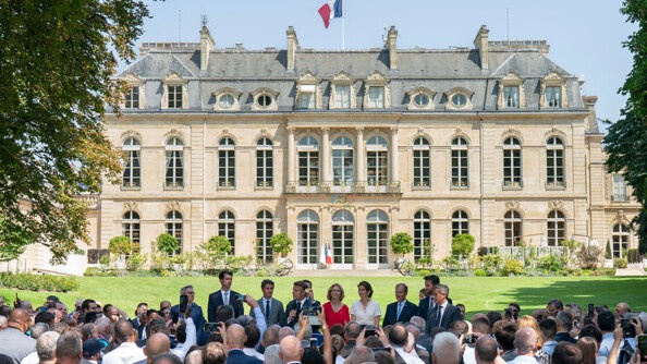 Une foule, que l'on aperçoit de dos, fait face au président de la République, Emmanuel Macron. Ce dernier est entouré de personnalités politiques. Derrière eux, se dresse le palais de l'Elysée.