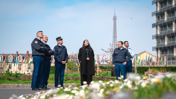 Cinq gendarmes et une femme en civil dans un jardin. EN arrière-plan à gauche plusieurs drapeaux de différents pays, en arrière-plan au fond la tour-eiffel