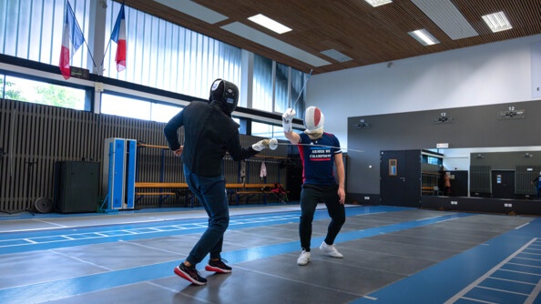 Deux escrimeurs s'affrontent sur une piste bleue et noire. le mur derrière eux est recouvert d'un miroir immense