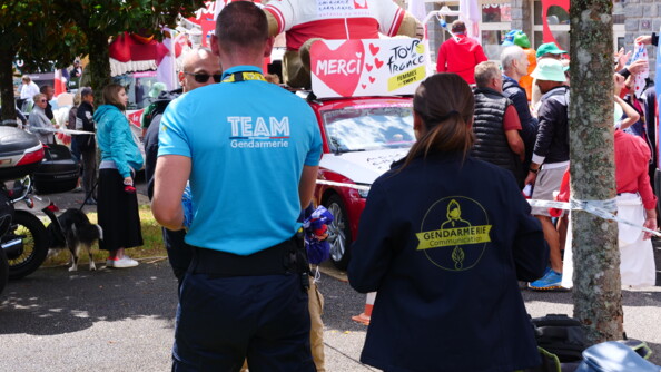 De dos marchant, à gauche un gendarme de la caravane publicitaire et à droite une gendarme portant la polaire communication de la gendarmerie, le long des  attractions du Tour de France féminin, dont une voiture rouge et blanche surmontée d'un ours géant avec lunettes de soleil et t-shirt rouge et blanc